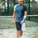 A man in sportswear (color - block shirt, split shorts, white cap) walks on a tennis court.