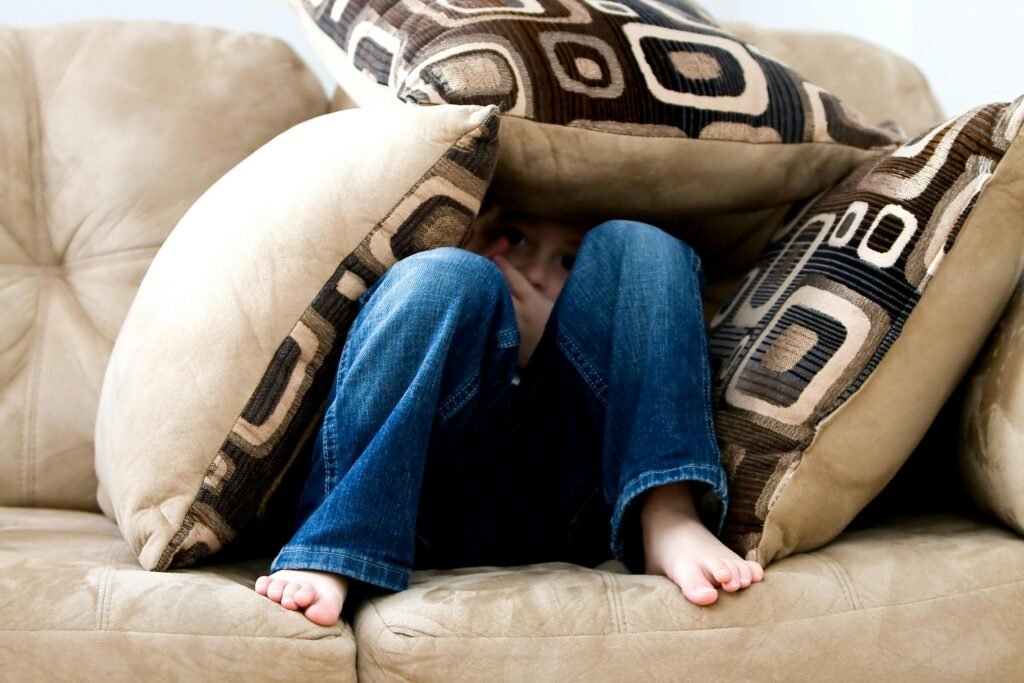 A child hides playfully under cushions on a beige sofa, peeking out with curiosity.